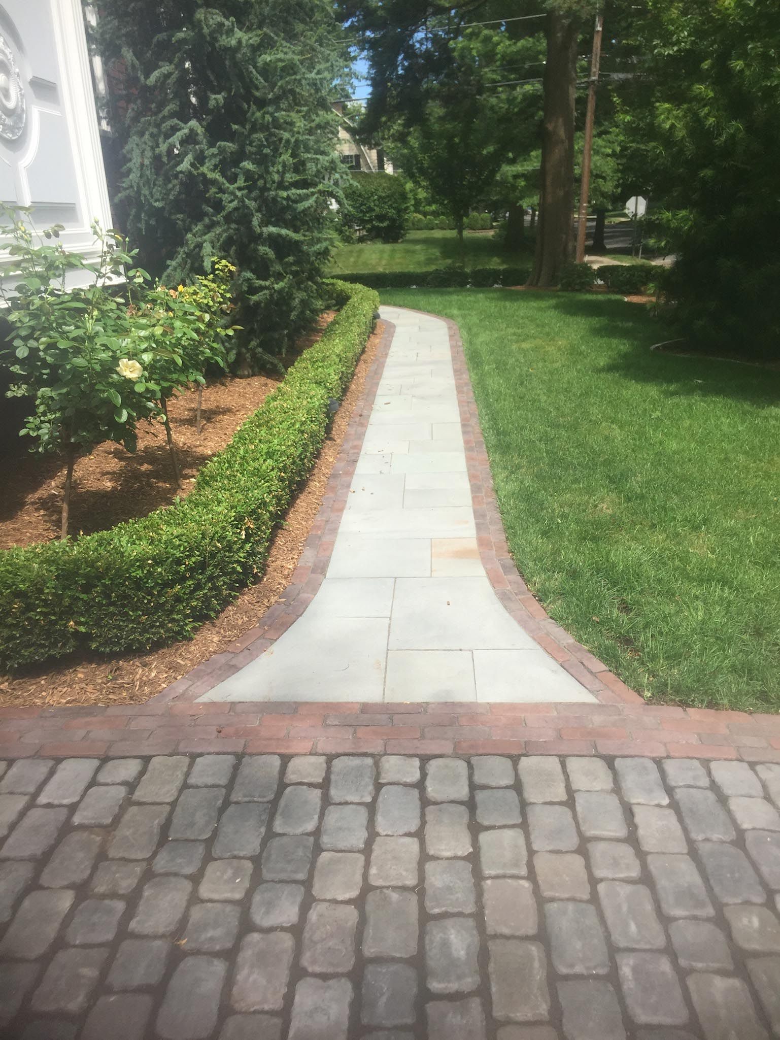 Brick walkway through green lawn beside a house and garden hedge, leading to a tree-lined yard
