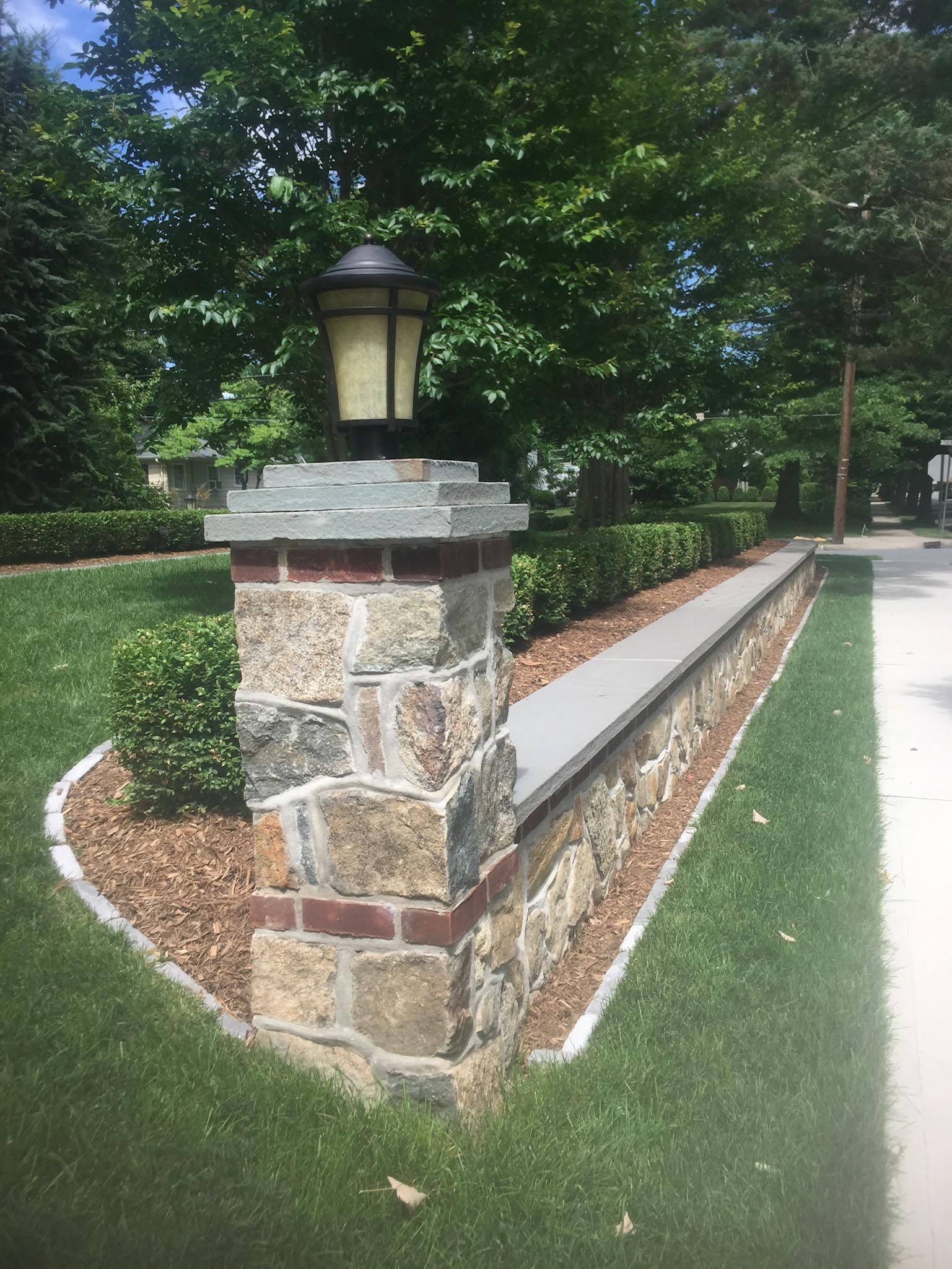 Stone garden wall with a lantern, bordered by greenery and a curved mulch bed along a sidewalk