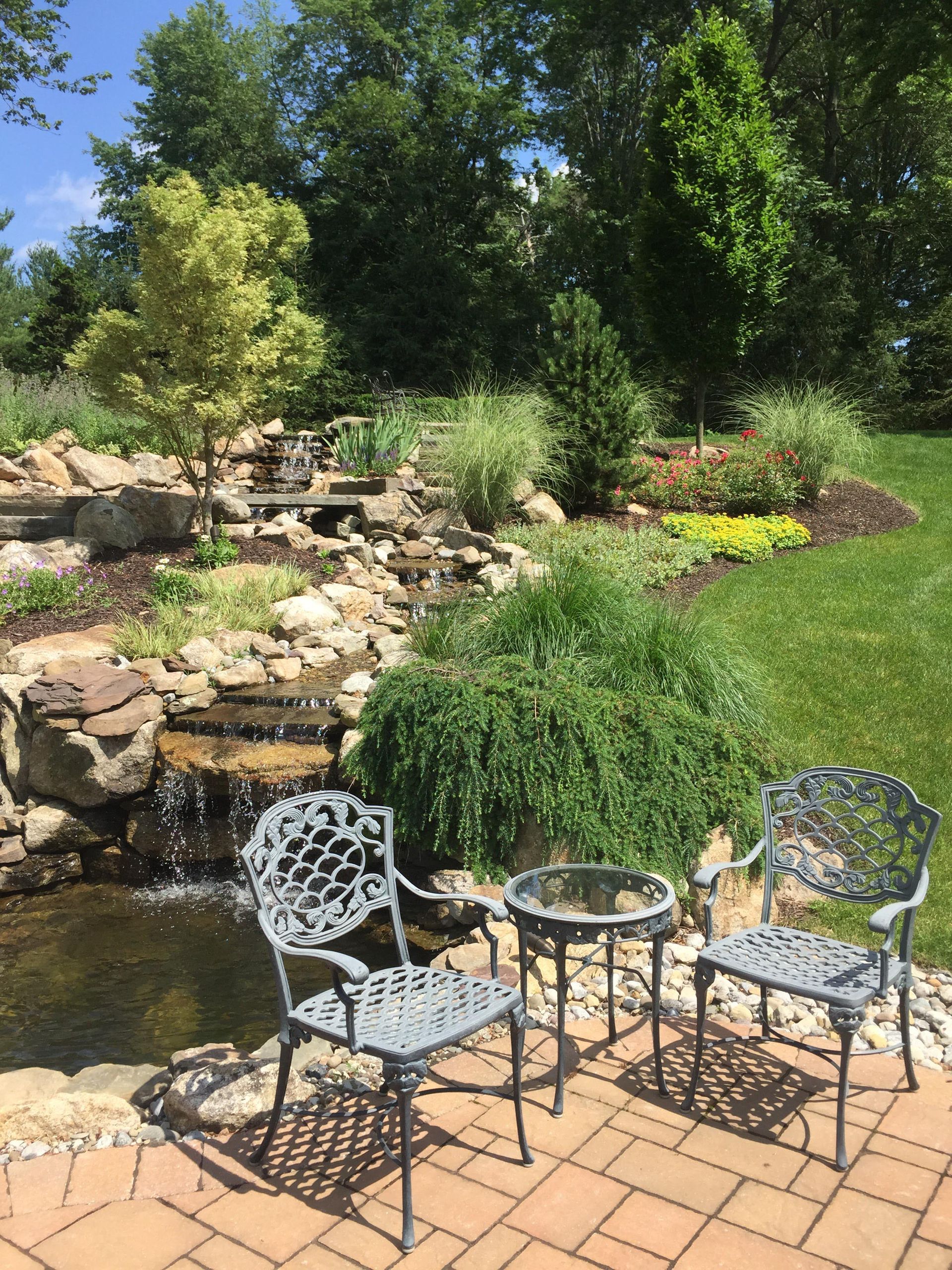 Patio chairs beside a small pond and waterfall in a lush garden with flowers and trees