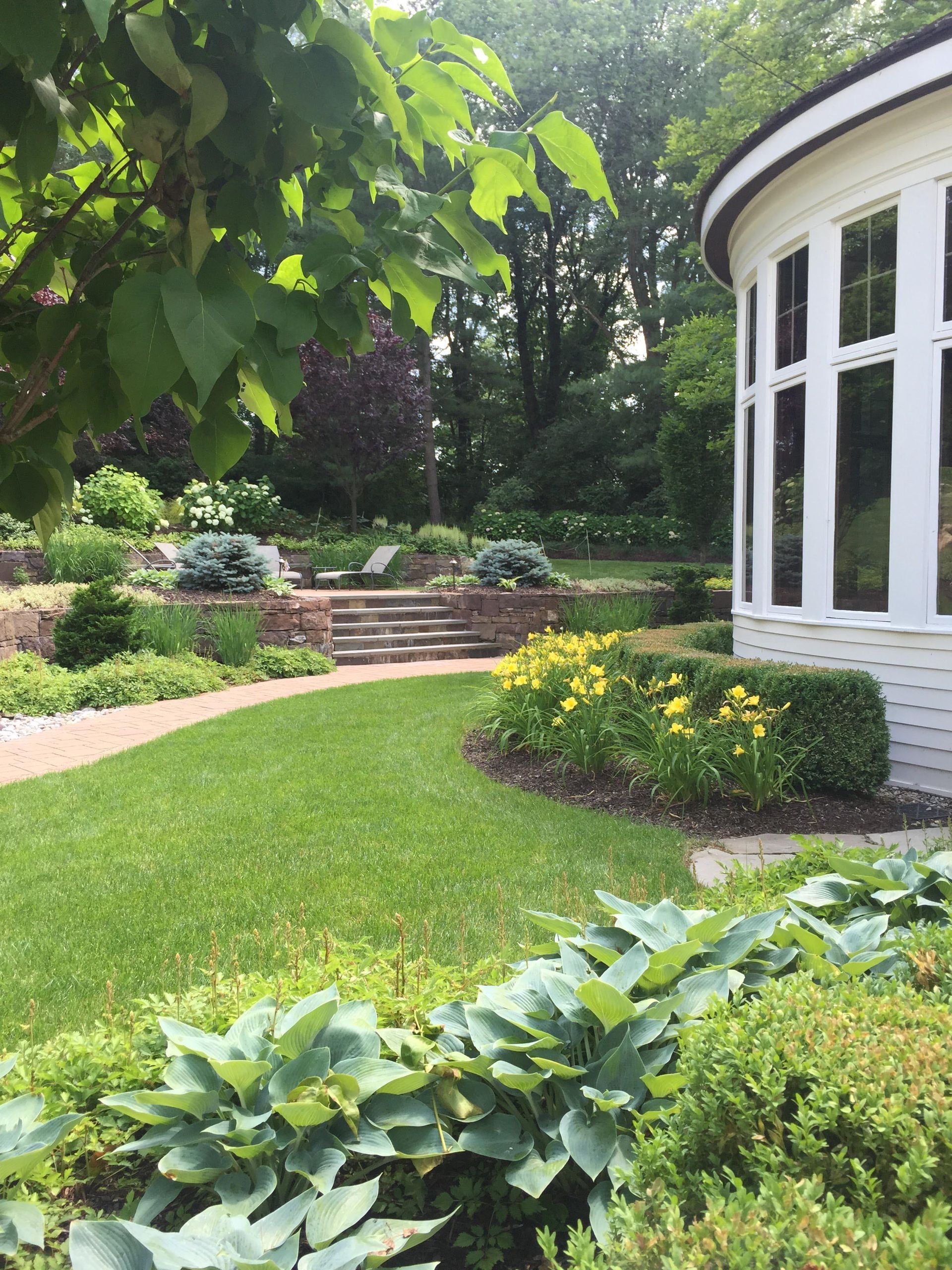 Curved white sunroom beside a lush green garden with stone steps and yellow flowers