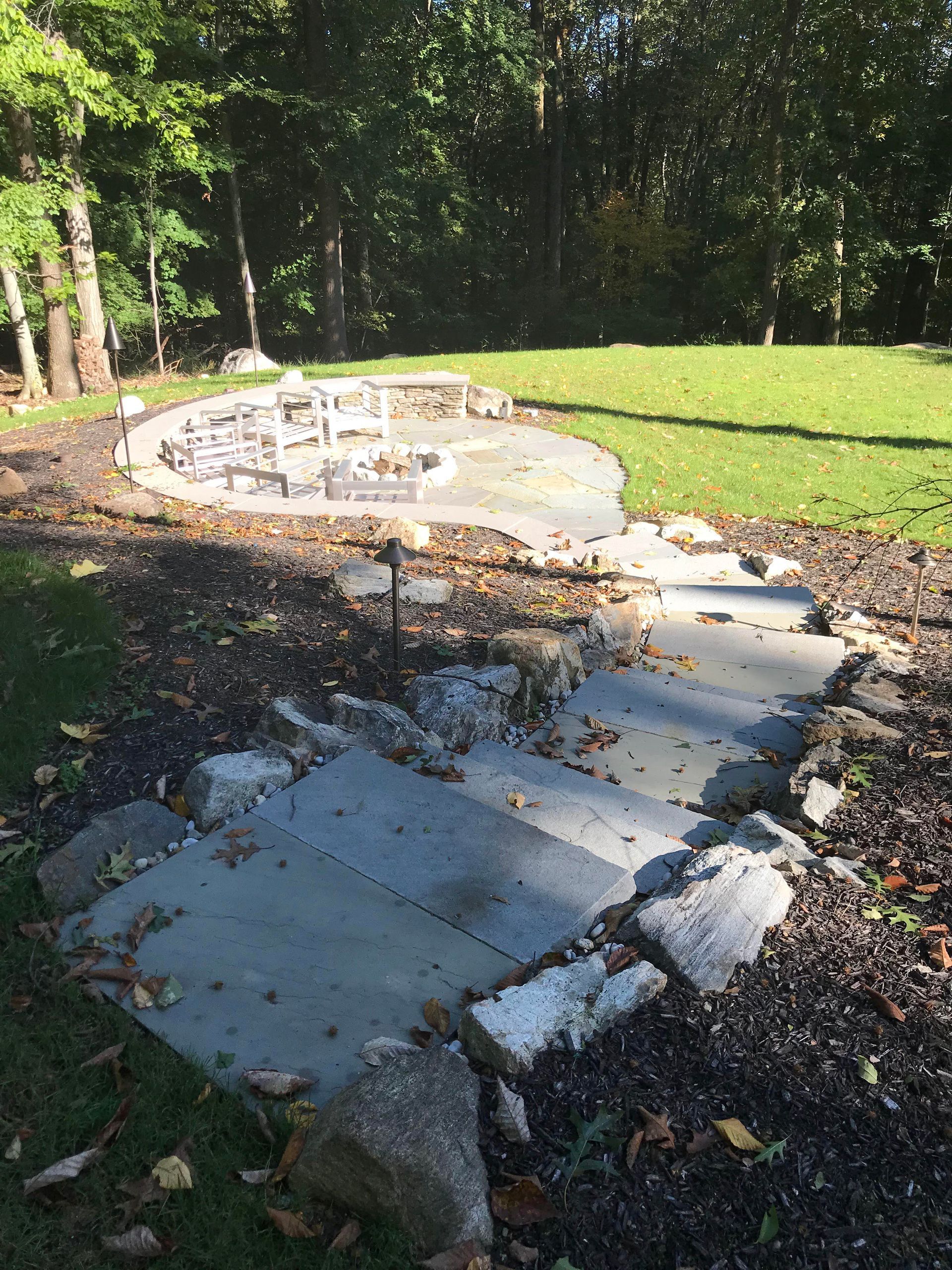 Stone steps lead to a circular patio in a shaded wooded backyard, bordered by grass and fallen leaves.