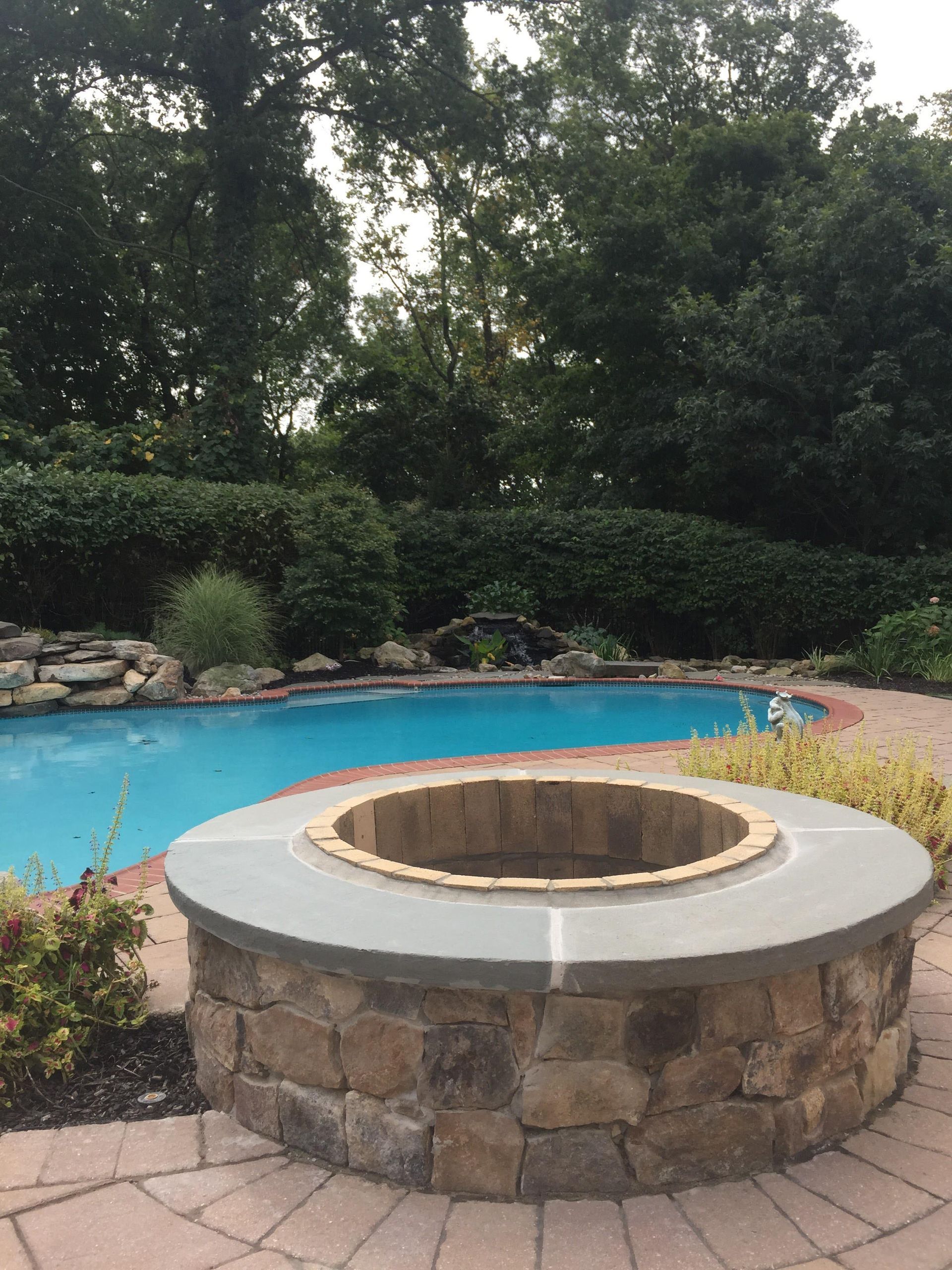 Stone fire pit beside a blue backyard pool, surrounded by trees and landscaped rocks.