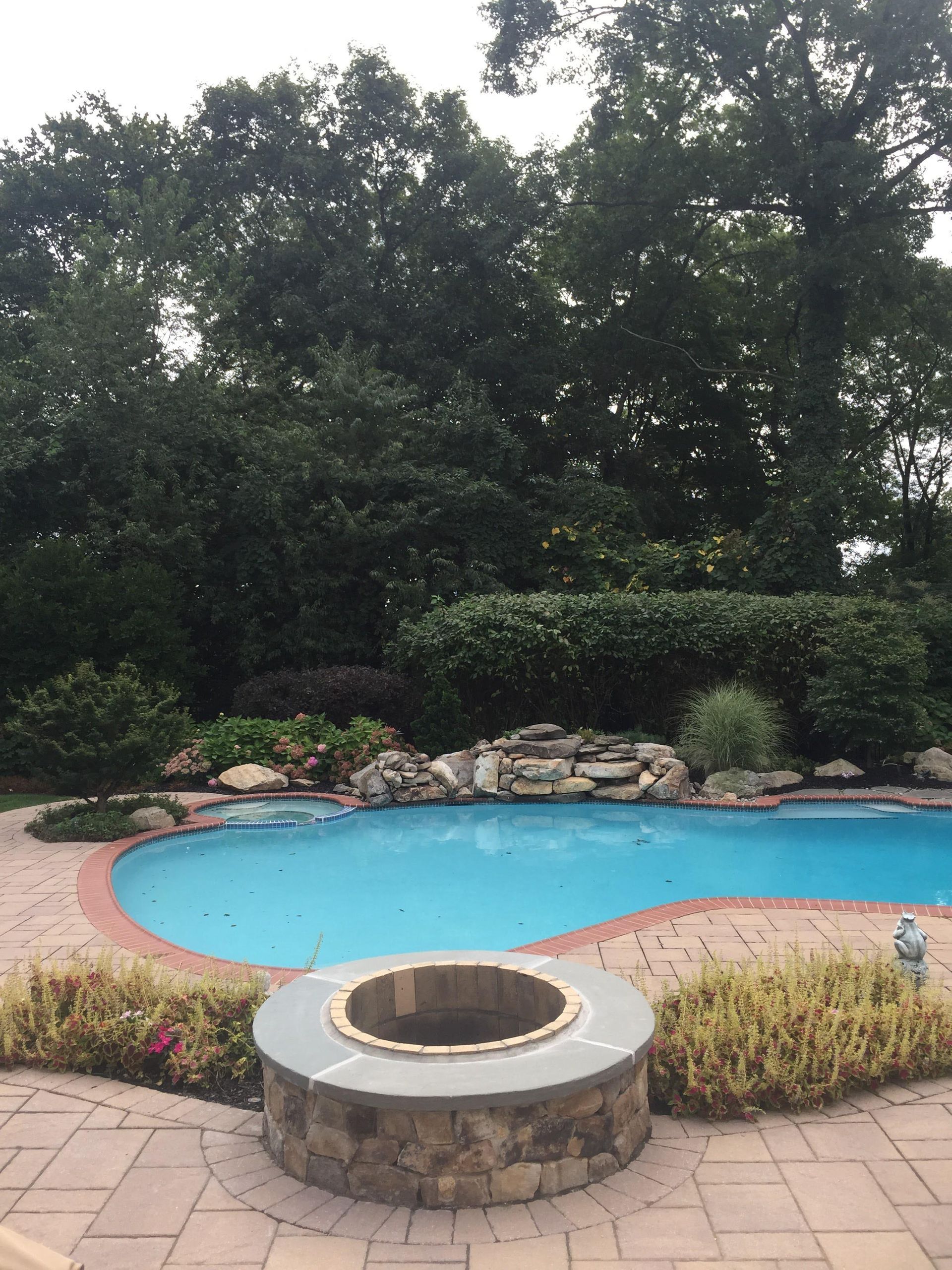Backyard pool with stone fire pit in front, surrounded by patio, rocks, and trees