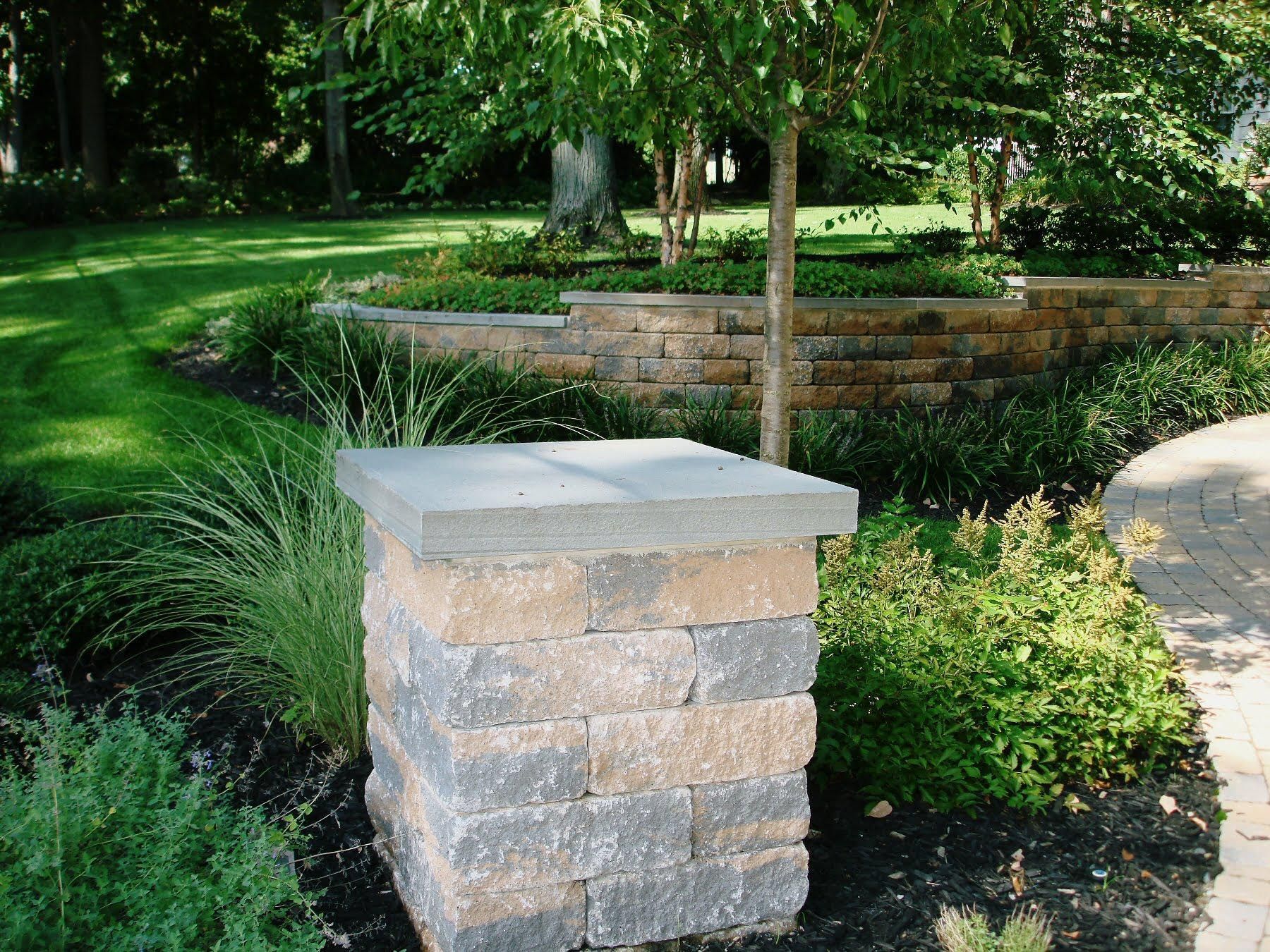 Stone pillar with a flat top in a landscaped garden with green shrubs and a brick retaining wall.