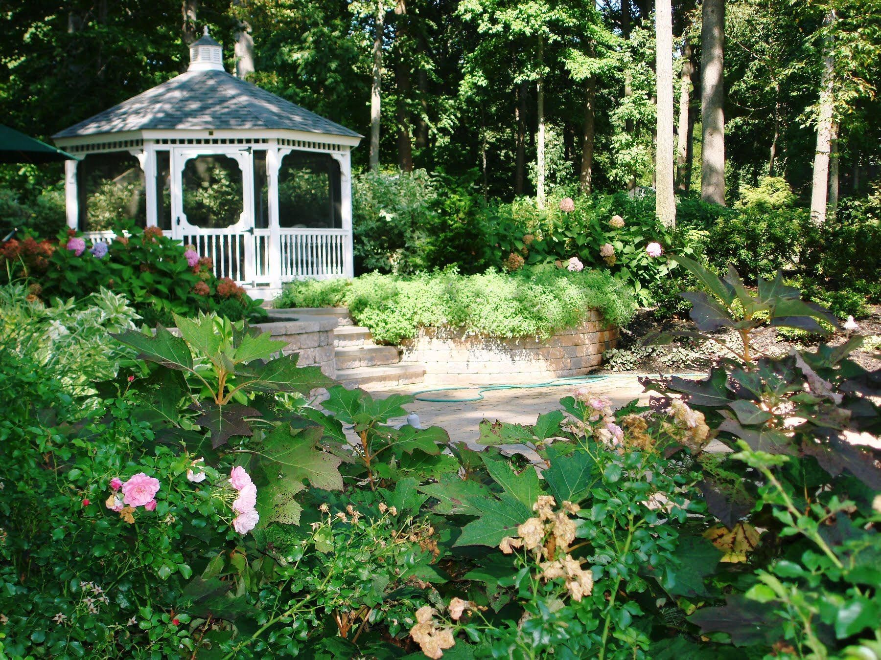 White gazebo in a lush garden with flowers, stone path, and tall trees in the background