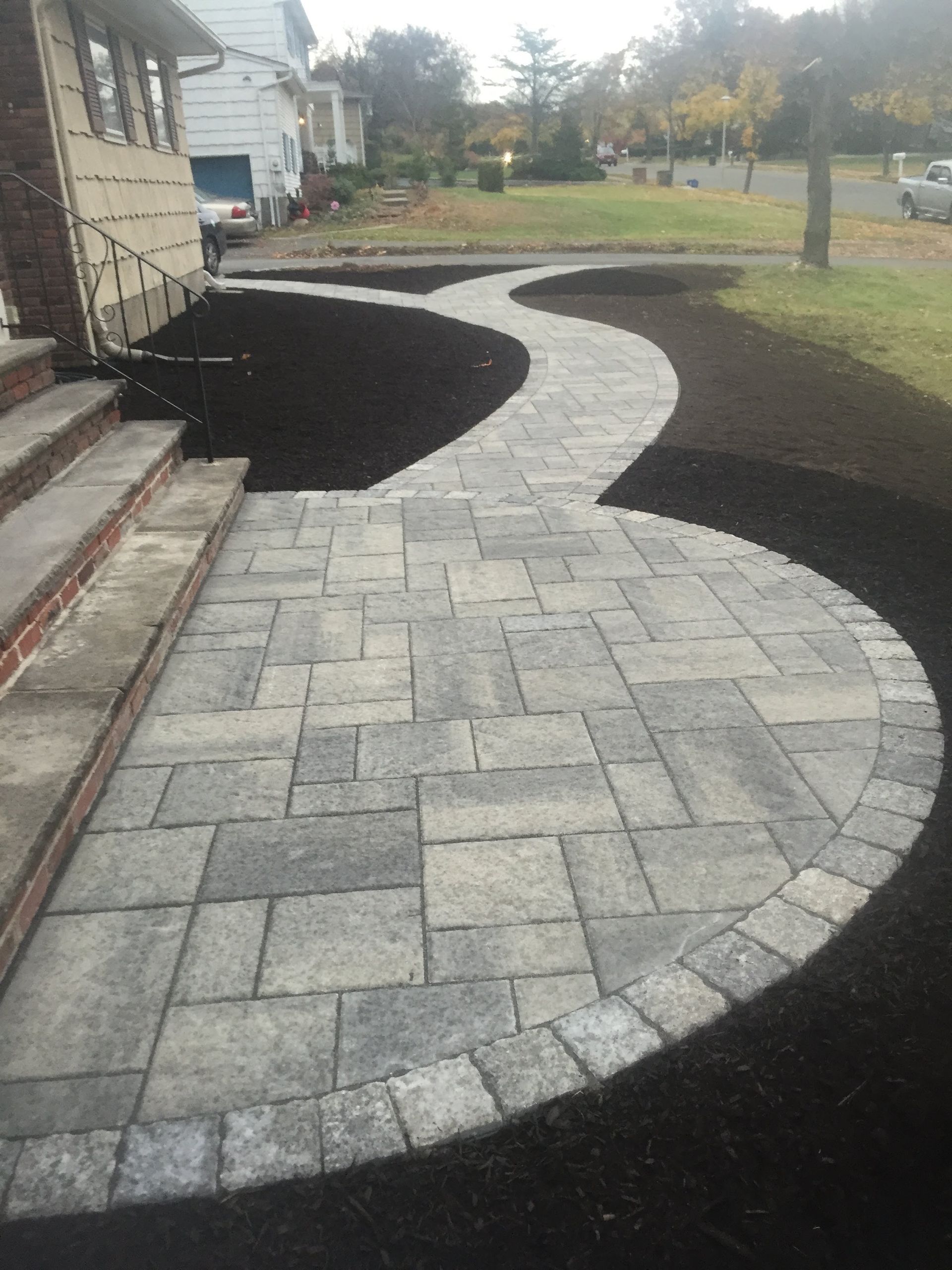 Curved stone paver walkway beside a house, bordered by dark mulch and grass in a residential yard.