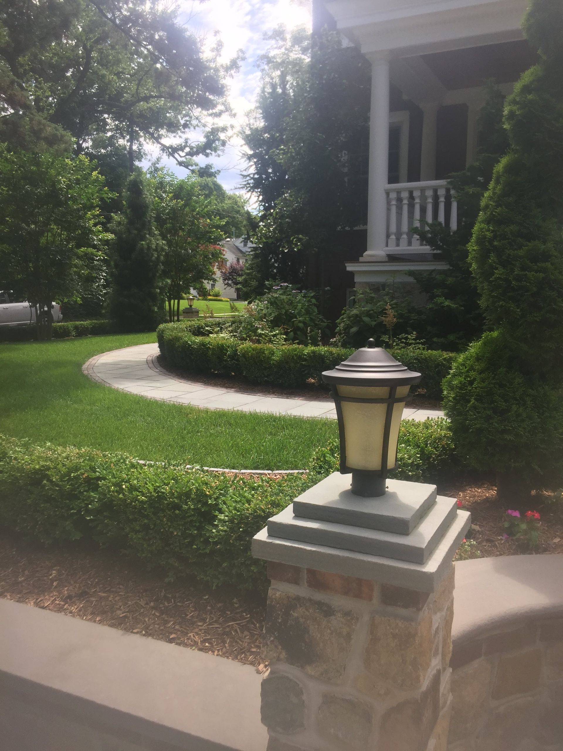 Front yard walkway, stone lamp post, manicured hedges, and a house porch in bright sunlight