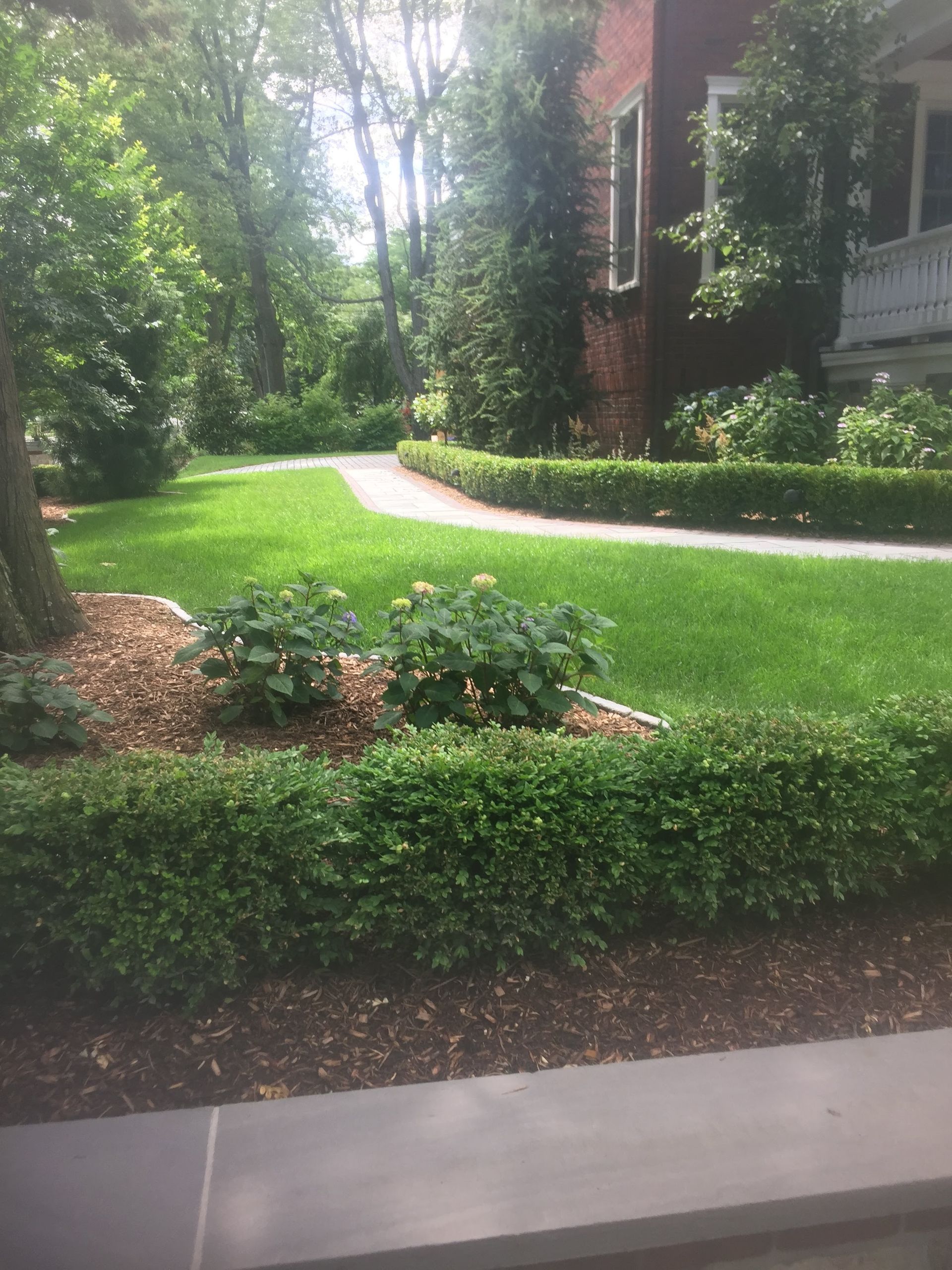 Manicured garden path beside a brick building, with green lawn, shrubs, and trees.