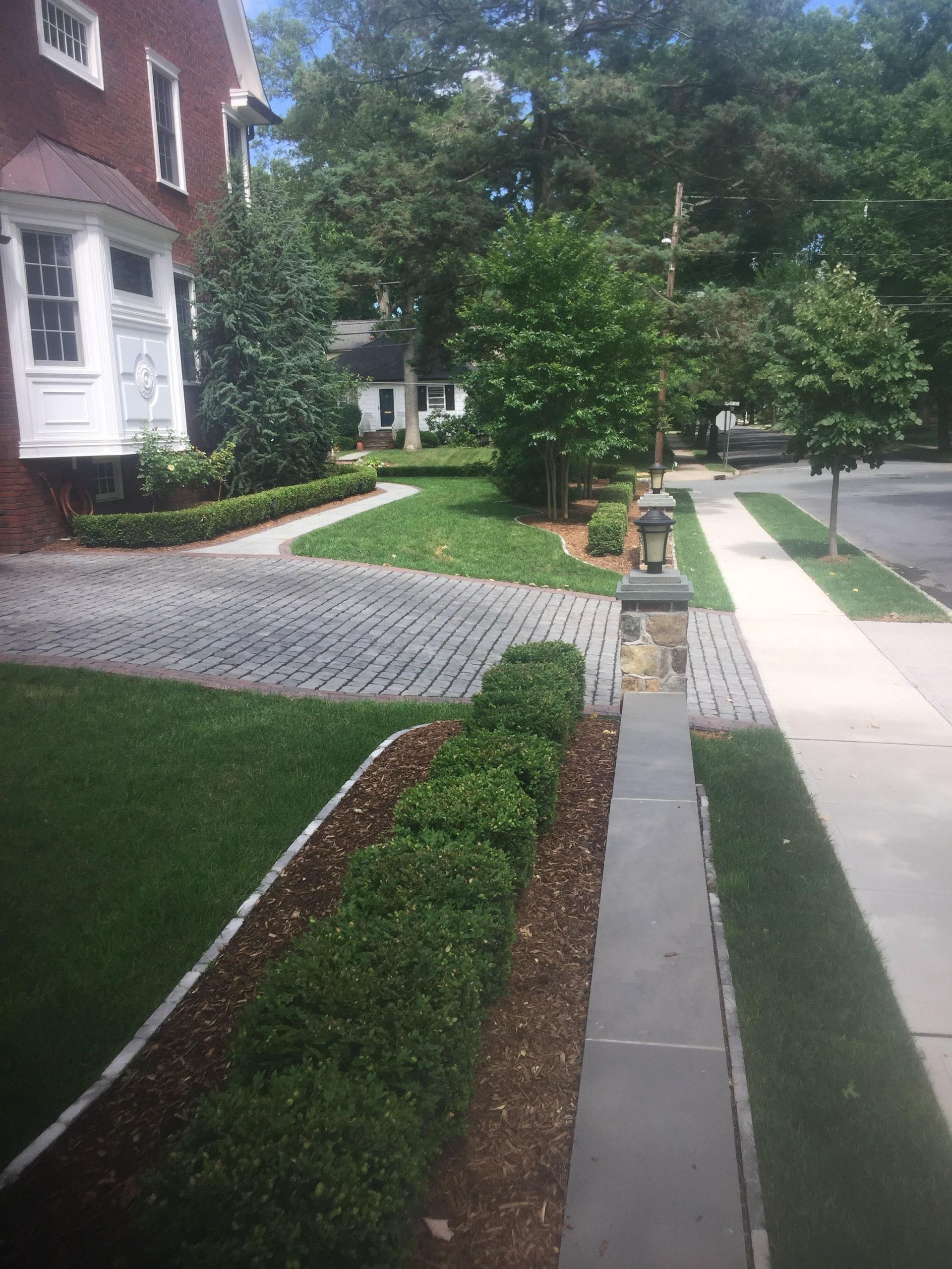 Front yard with a paved walkway, trimmed hedges, green lawn, and brick houses on a quiet residential street