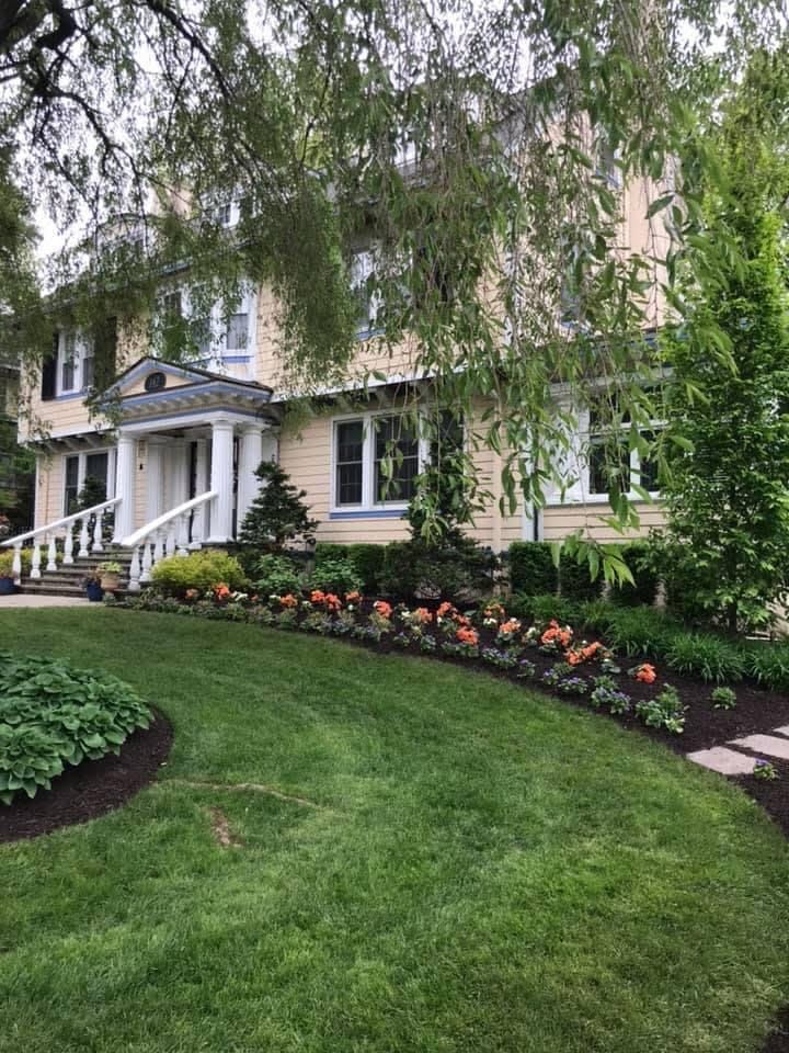 White house with porch, flowering garden beds, and lush green lawn under overhanging trees