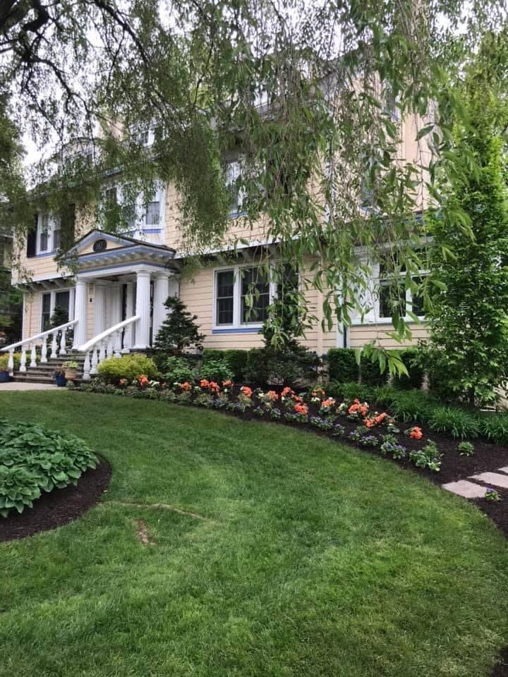White house with porch, lush green lawn, and flower beds under hanging tree branches