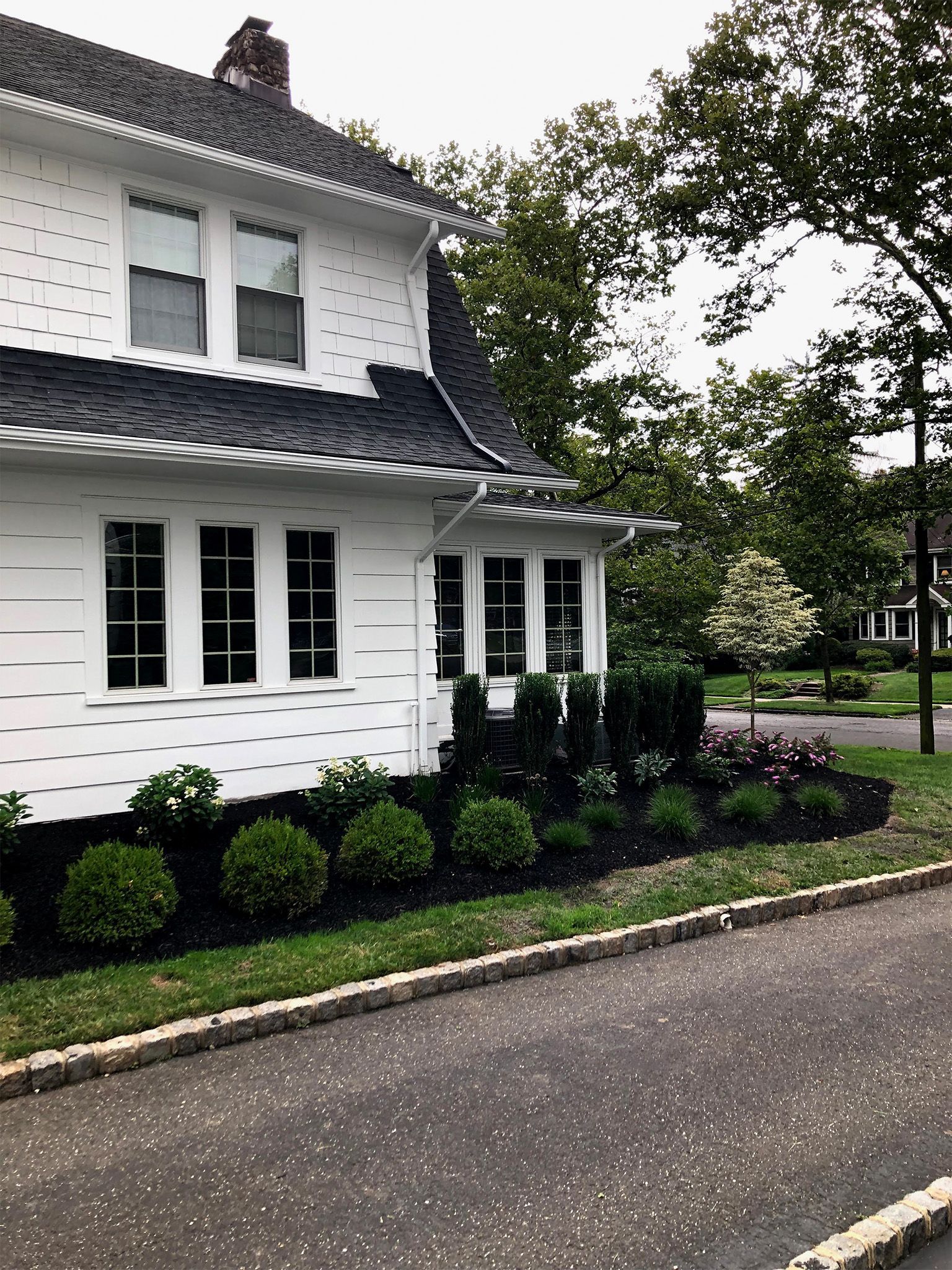 White two-story house with dark roof, trimmed shrubs, and a paved driveway under overcast skies