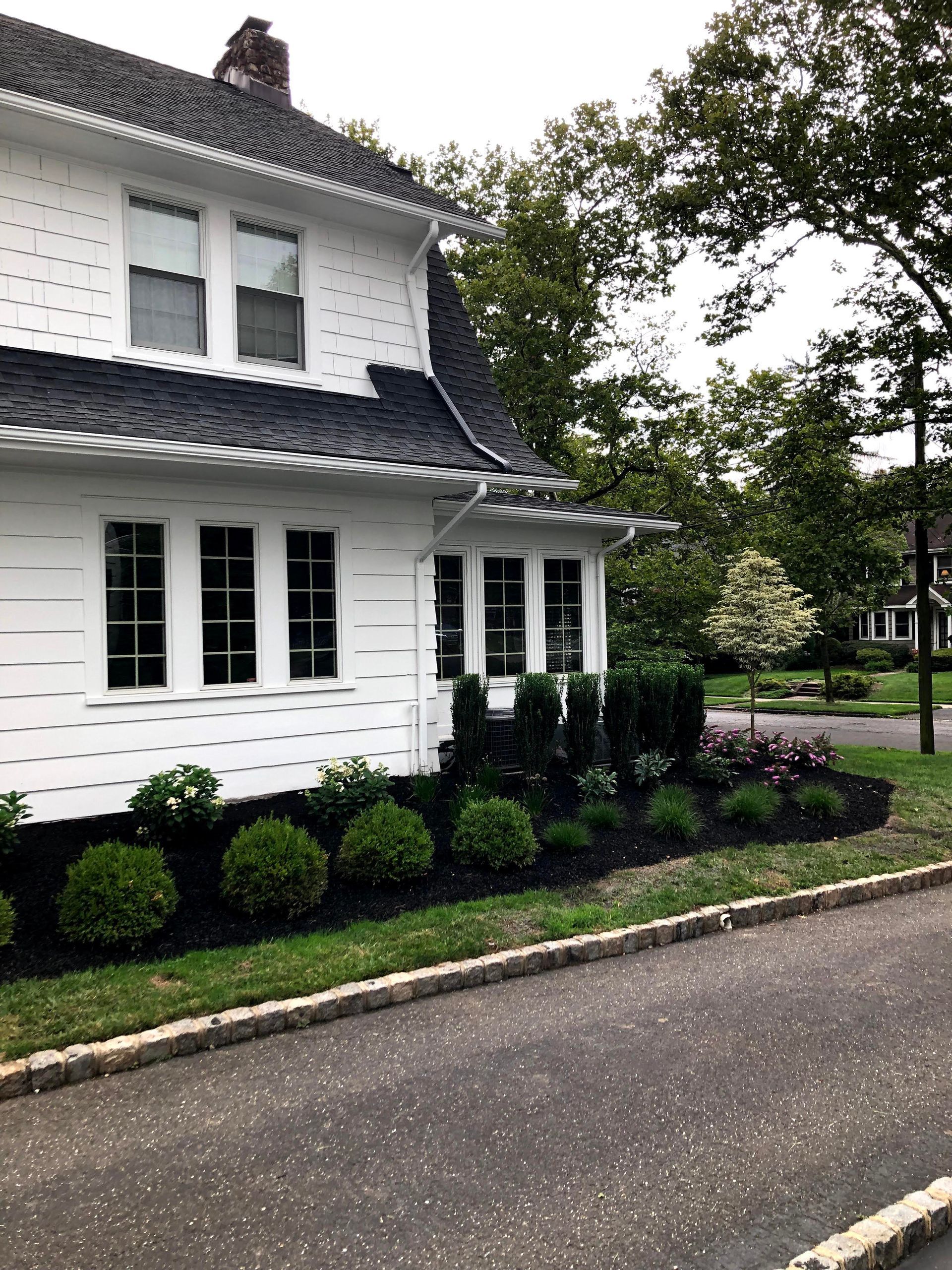 White two-story house with dark roof, trimmed shrubs, and a sidewalk along the street