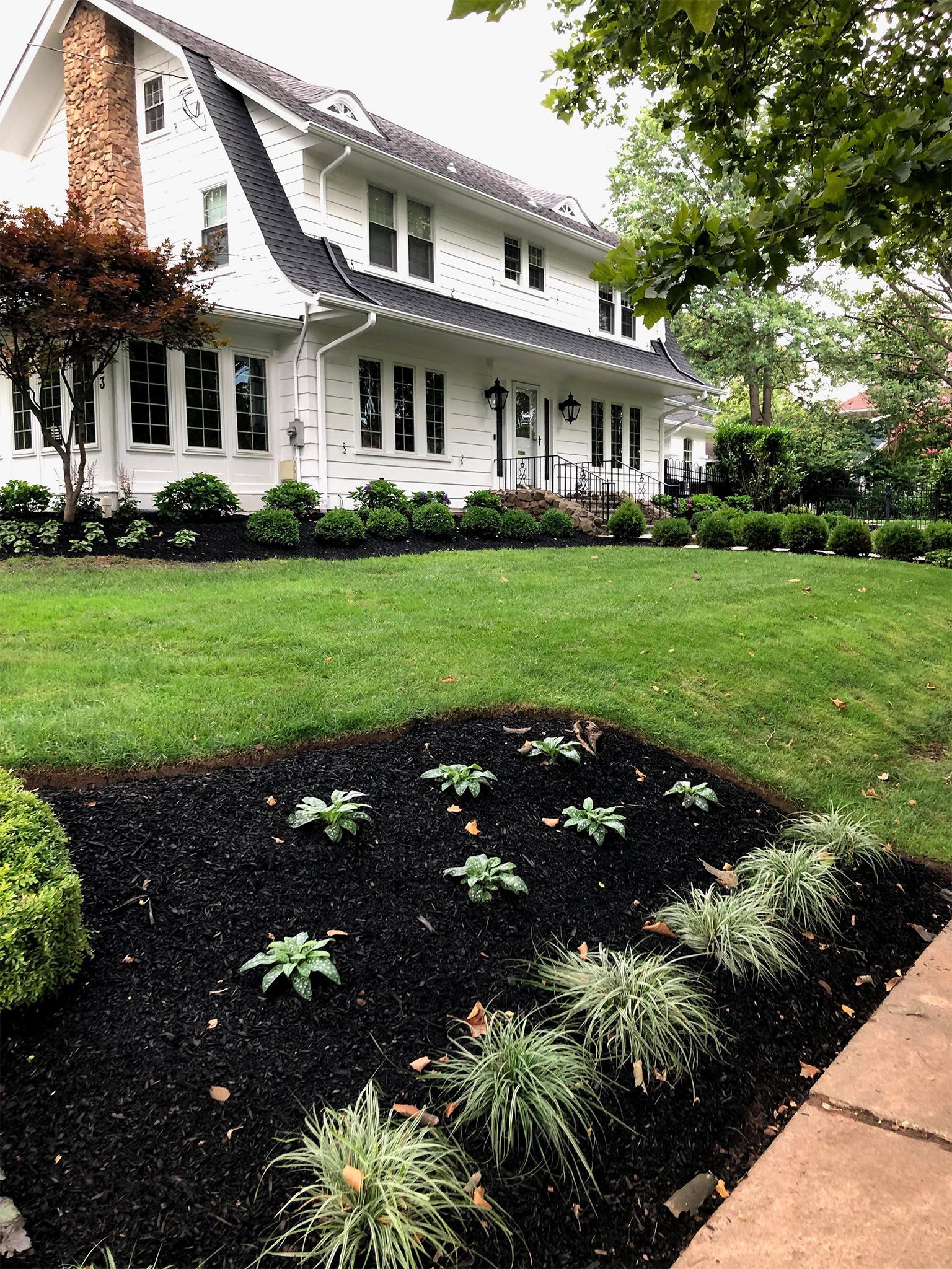 White house with black shutters beside a green lawn and flower bed under overcast skies