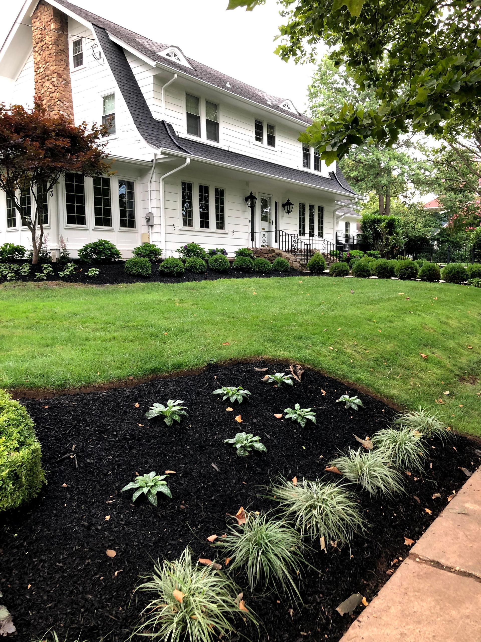 White two-story house with dark roof, green lawn, and mulched garden beds by a sidewalk