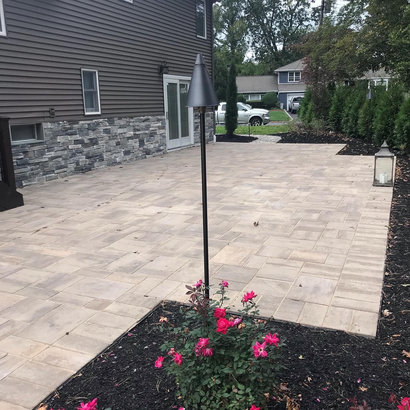 Backyard stone patio beside a house, with a slim lamp post and pink flowers in the foreground.