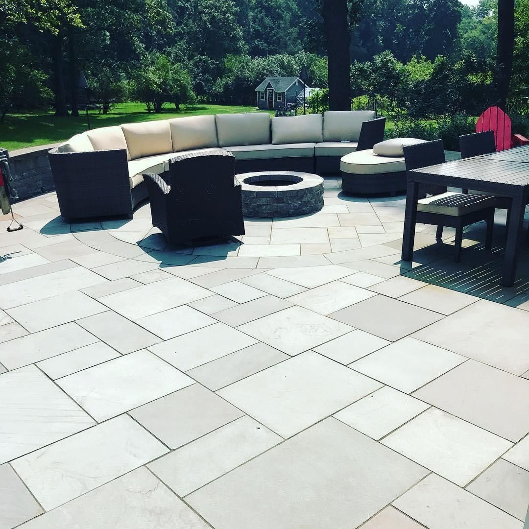 Outdoor patio with white pavers, curved beige sectional, fire pit, and black tables in a sunny backyard