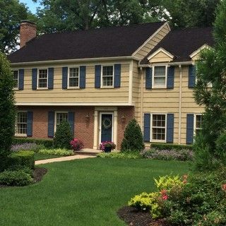 Suburban two-story beige house with dark roof, blue shutters, brick columns, and a manicured green lawn