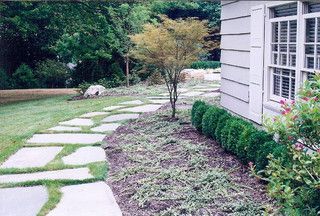 Stone path beside a house, leading through a green garden with shrubs and trees.
