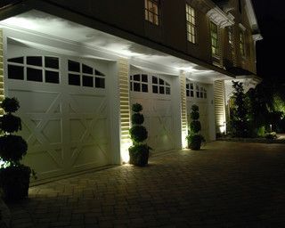 Lit suburban garage with white doors, potted topiary, and driveway at night