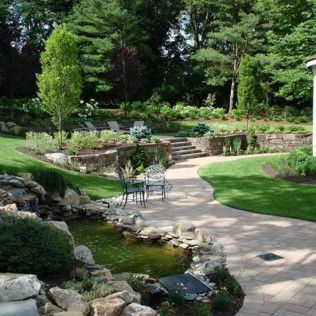 Landscaped garden with stone path, small stream, patio chairs, and lush green trees and shrubs