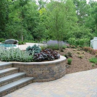 Stone patio steps leading to a curved raised garden bed with shrubs and trees in a landscaped yard