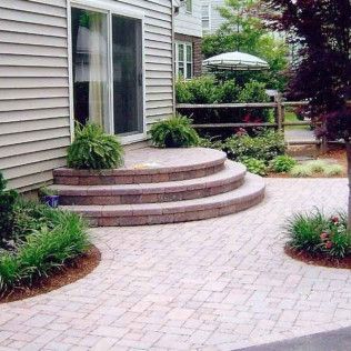 Curved brick walkway leading to a small raised patio with steps beside a house and garden plants