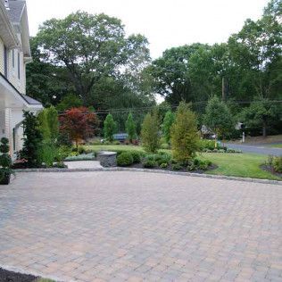 Brick-paved driveway beside a house, with landscaped shrubs, trees, and a quiet street in the background.