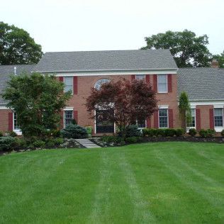Brick house with gray roof, red shutters, and a manicured green lawn bordered by trees.