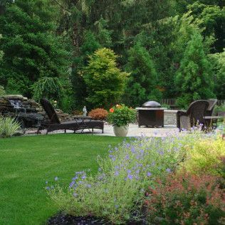 Lush backyard patio with wicker chairs, fire pit, and colorful flowers surrounded by green trees