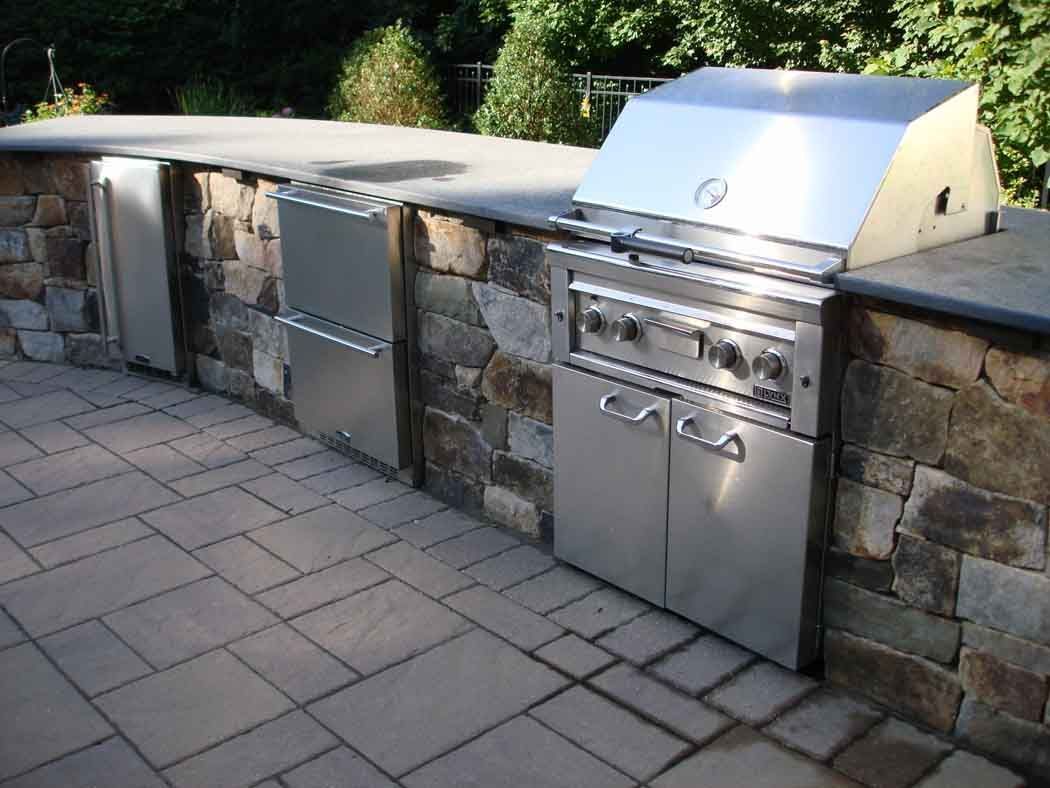 Stone outdoor kitchen with built-in grill, sink, and storage cabinets on a paved patio