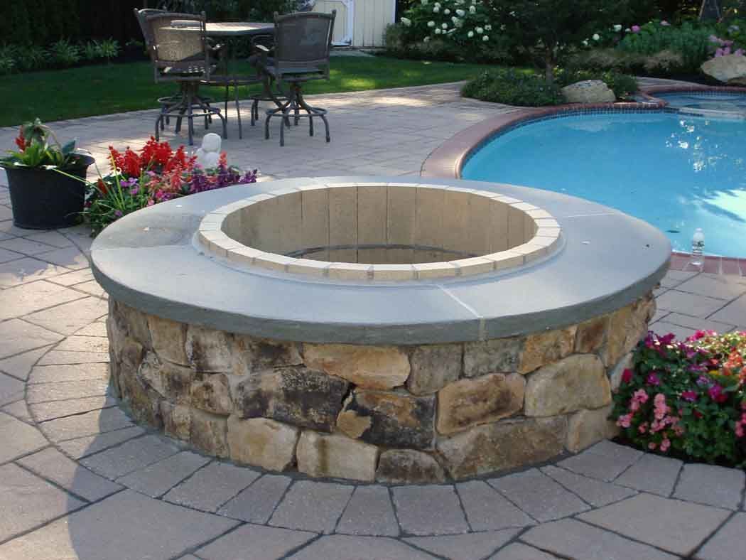 Stone fire pit beside a swimming pool, surrounded by patio pavers and flowers.