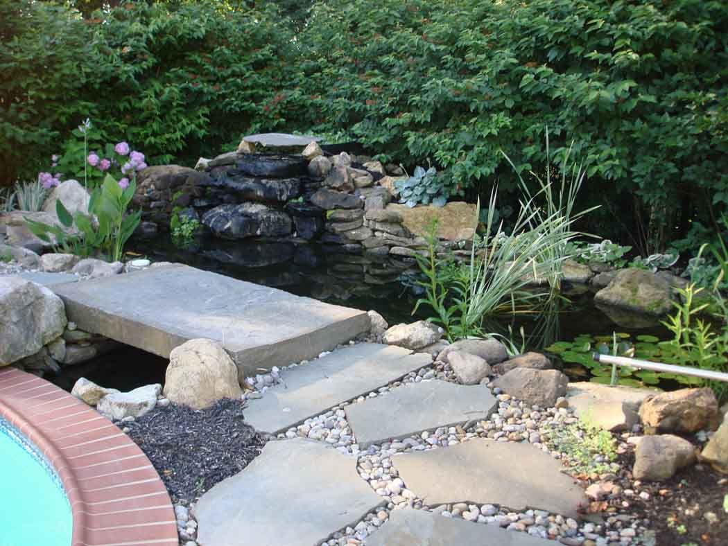 Stone patio with a small pond, stepping stones, and lush greenery in a garden