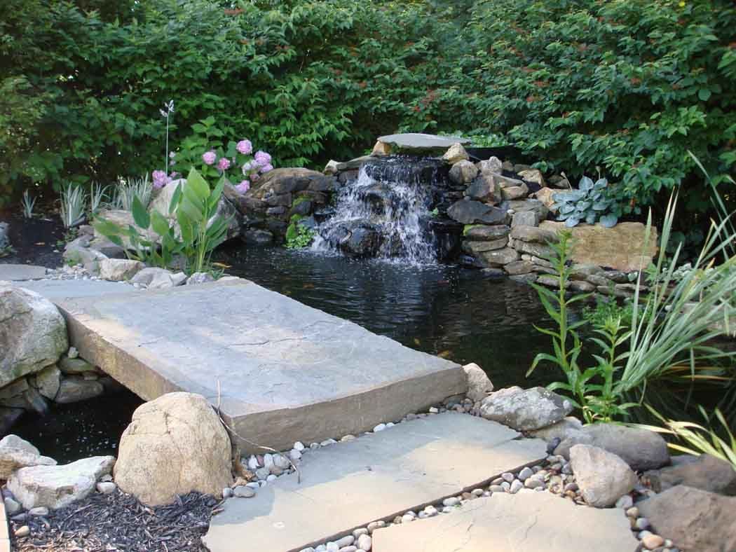 Garden pond with small waterfall, stone bridge, and flowers surrounded by rocks and greenery