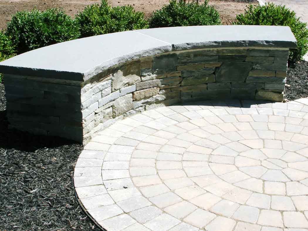 Curved stone bench beside a light paver patio with shrubs in the background