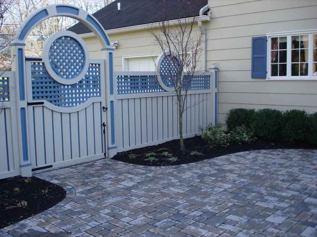 White picket fence gate with blue circular trellis beside a beige house and stone-paver driveway