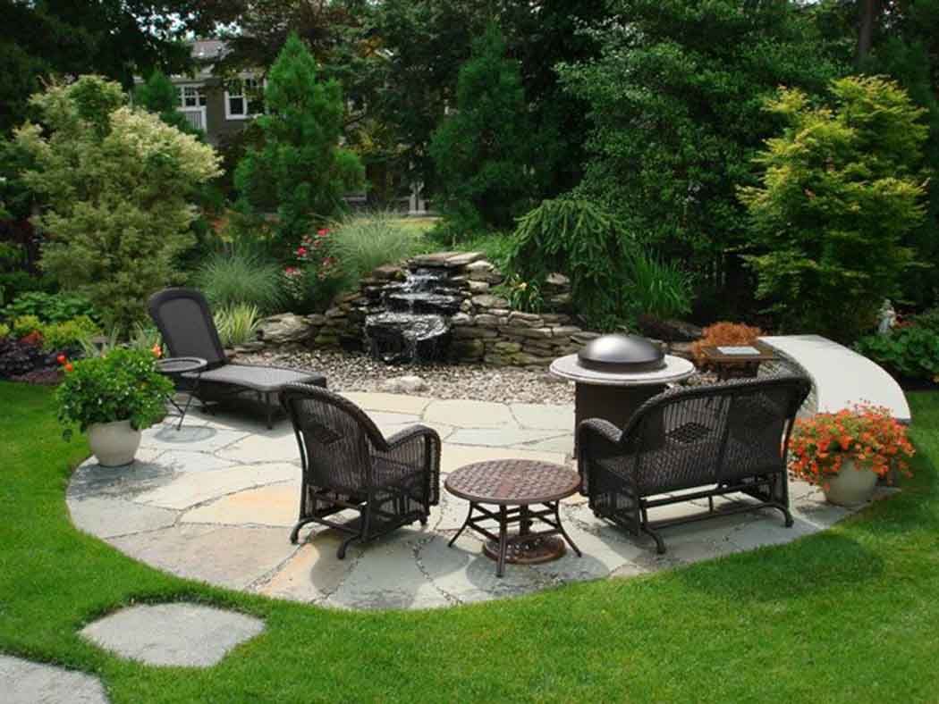 Backyard patio with wicker chairs around a fire pit, stone seating area, and lush green landscaping