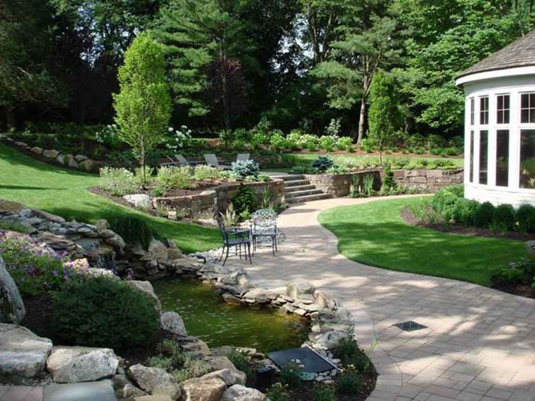 Lush garden with stone path, small pond, patio chairs, and a white gazebo in the background