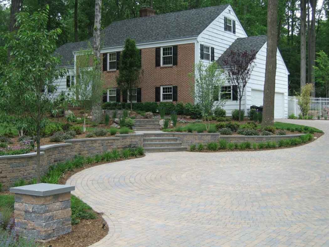 Curved stone driveway leading to a brown-and-white house with landscaped garden and trees