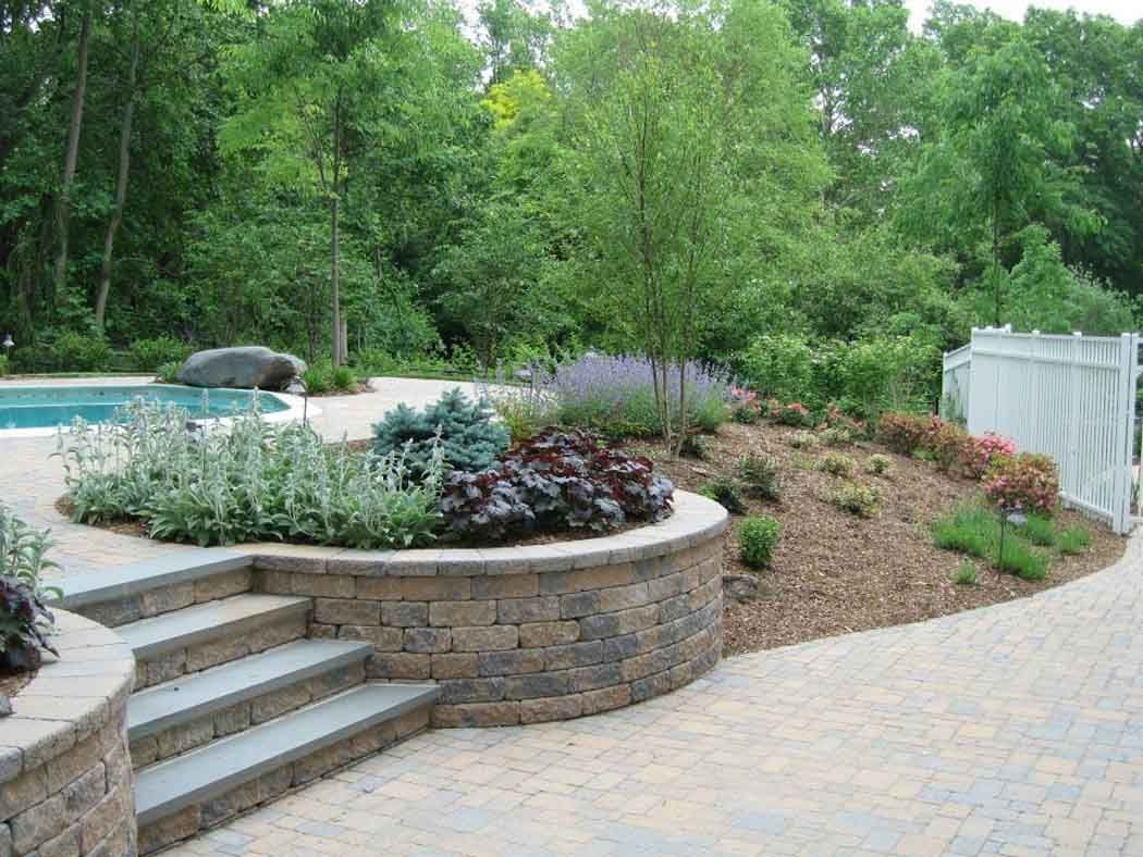 Curved stone patio with steps, landscaped flower bed, and paved walkway in a green backyard