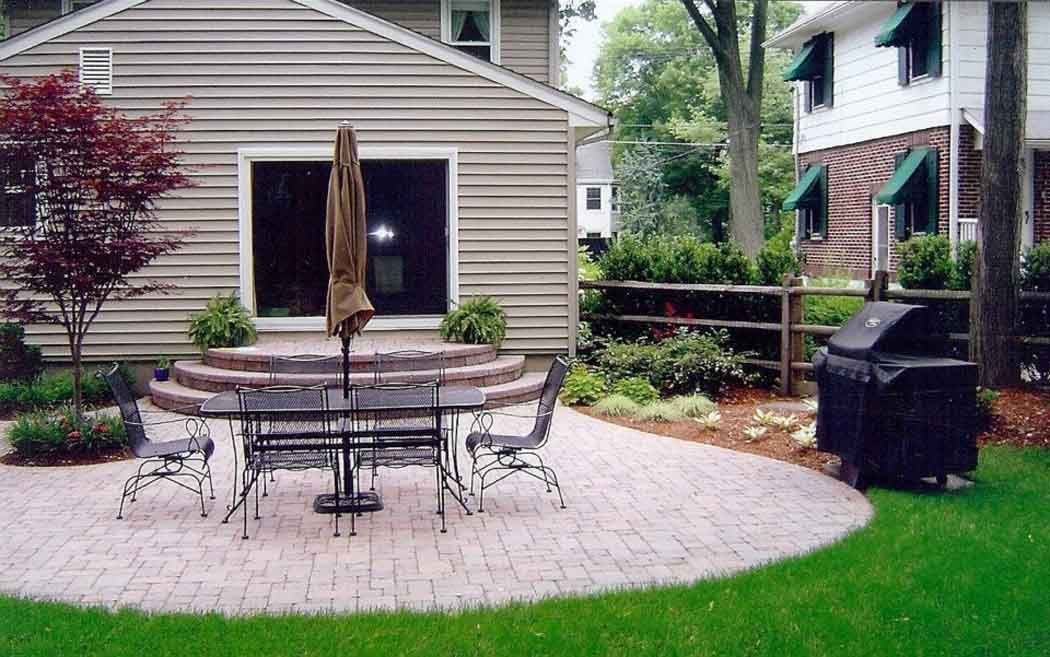 Backyard patio with round table, closed umbrella, grill, and beige house with sliding door