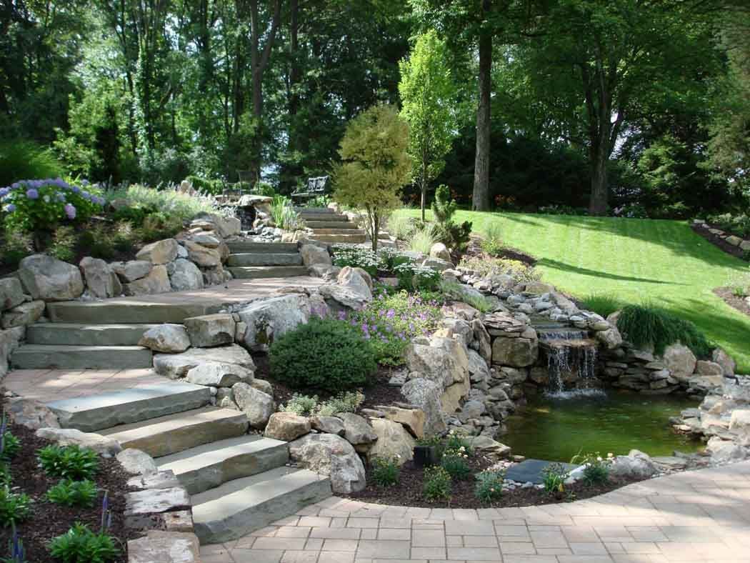 Stone steps beside a small garden pond with rocks, flowers, and lush green trees
