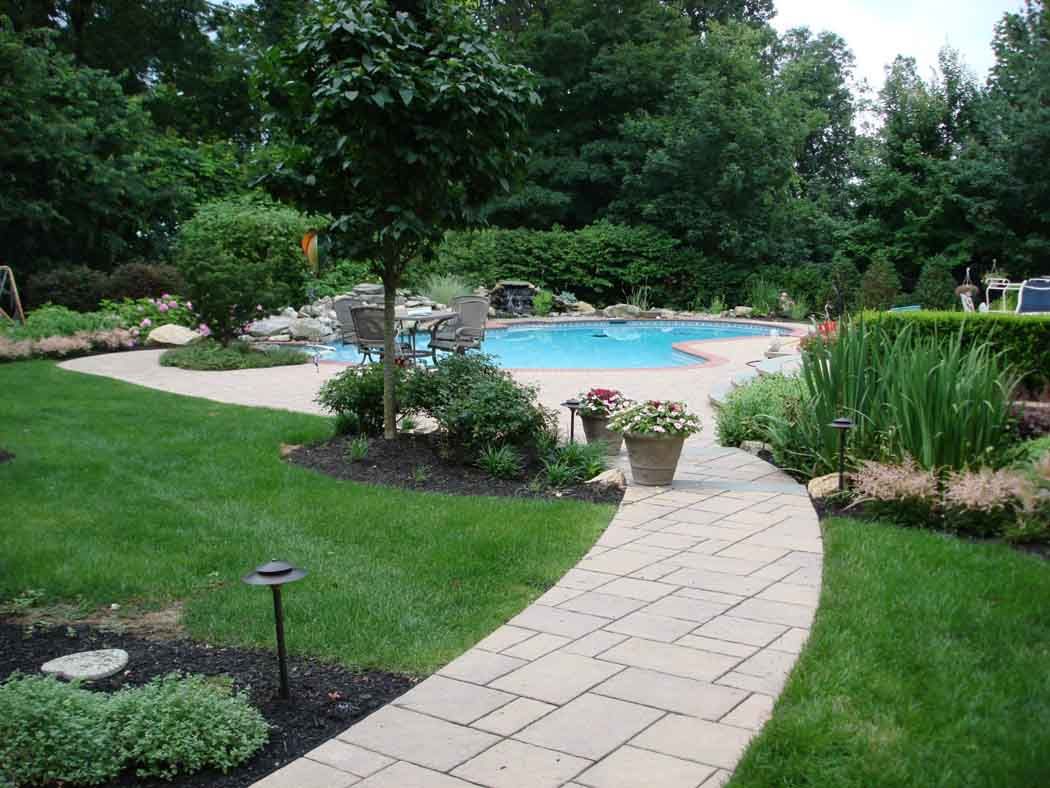 Backyard garden with curved stone path leading to a blue swimming pool surrounded by lush greenery