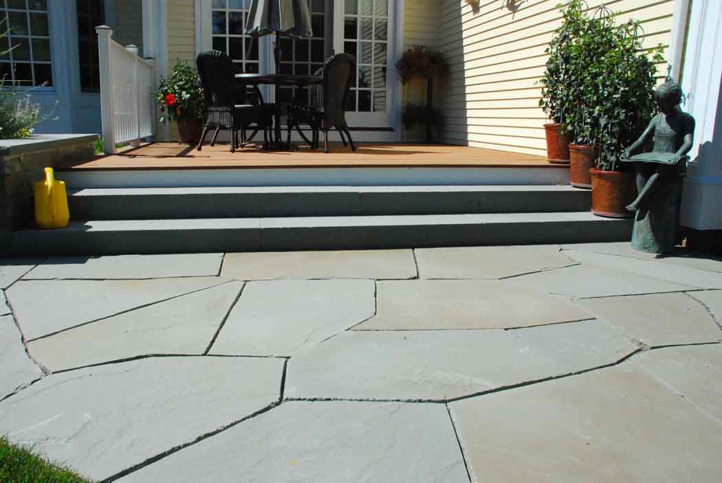 Stone patio leading to a backyard deck with steps, chairs, and potted plants