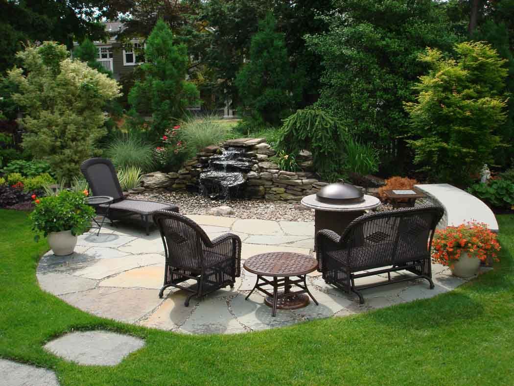 Backyard patio seating area with wicker chairs, fire pit, and garden waterfall surrounded by greenery