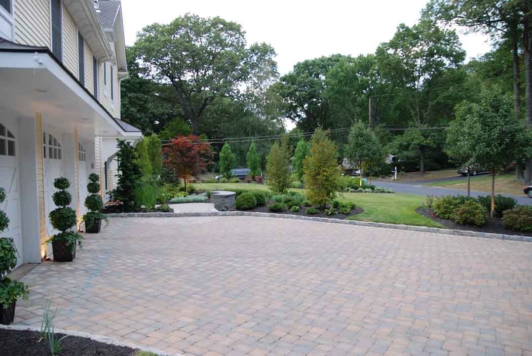 Brick driveway beside a white house, with landscaped garden and trees in the background