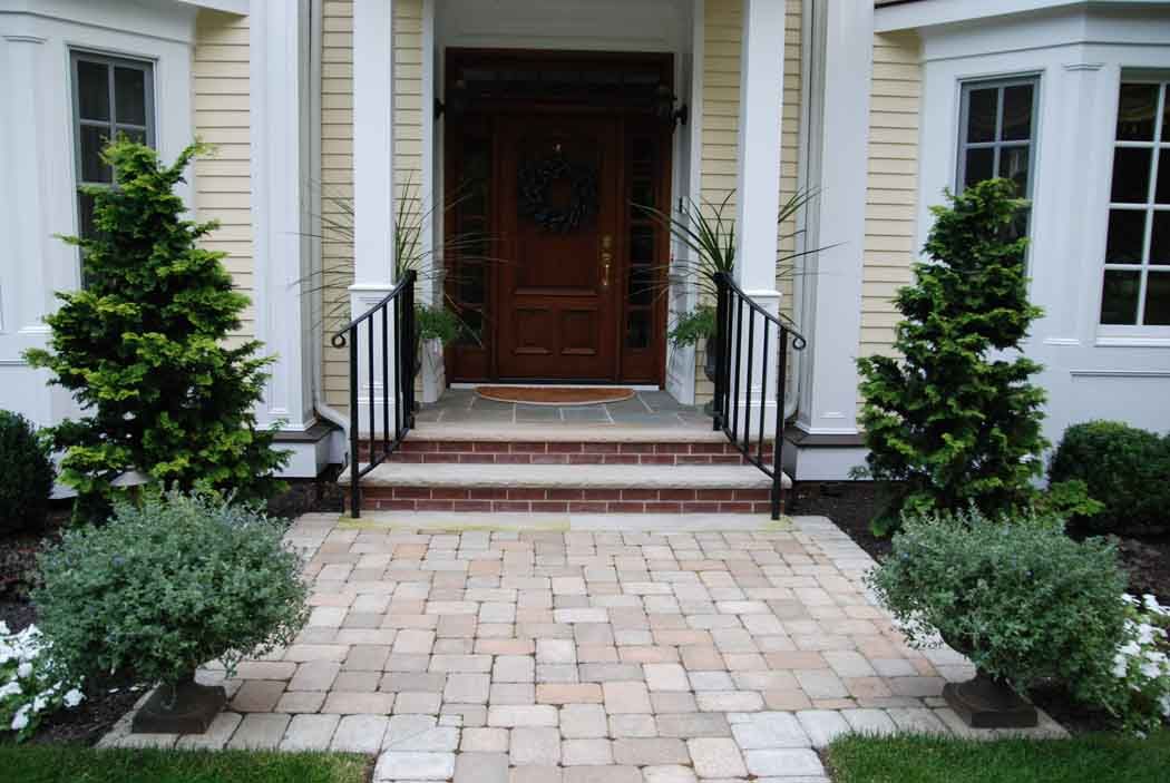 Brick walkway leading to a front porch with dark wooden door, white siding, and manicured shrubs