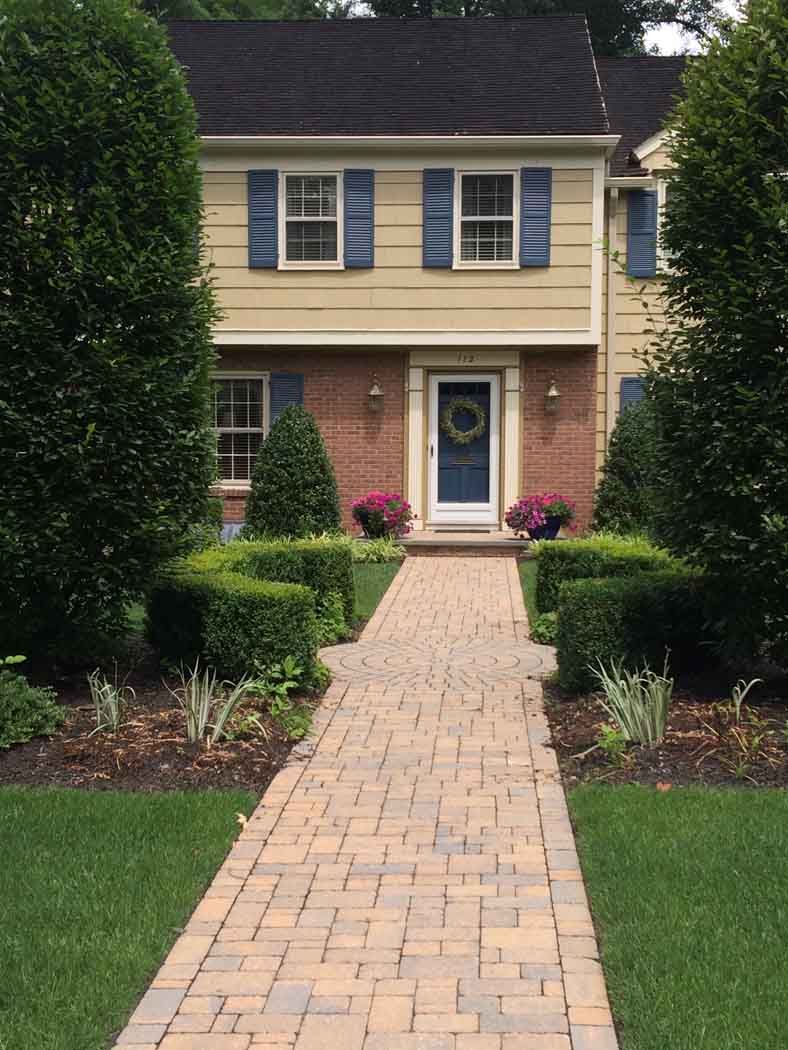 Brick path leading to a two-story yellow house with blue shutters and a front door framed by shrubs