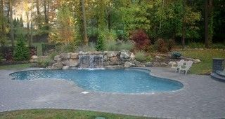 Backyard pool with a rock waterfall, blue water, and stone patio surrounded by trees.