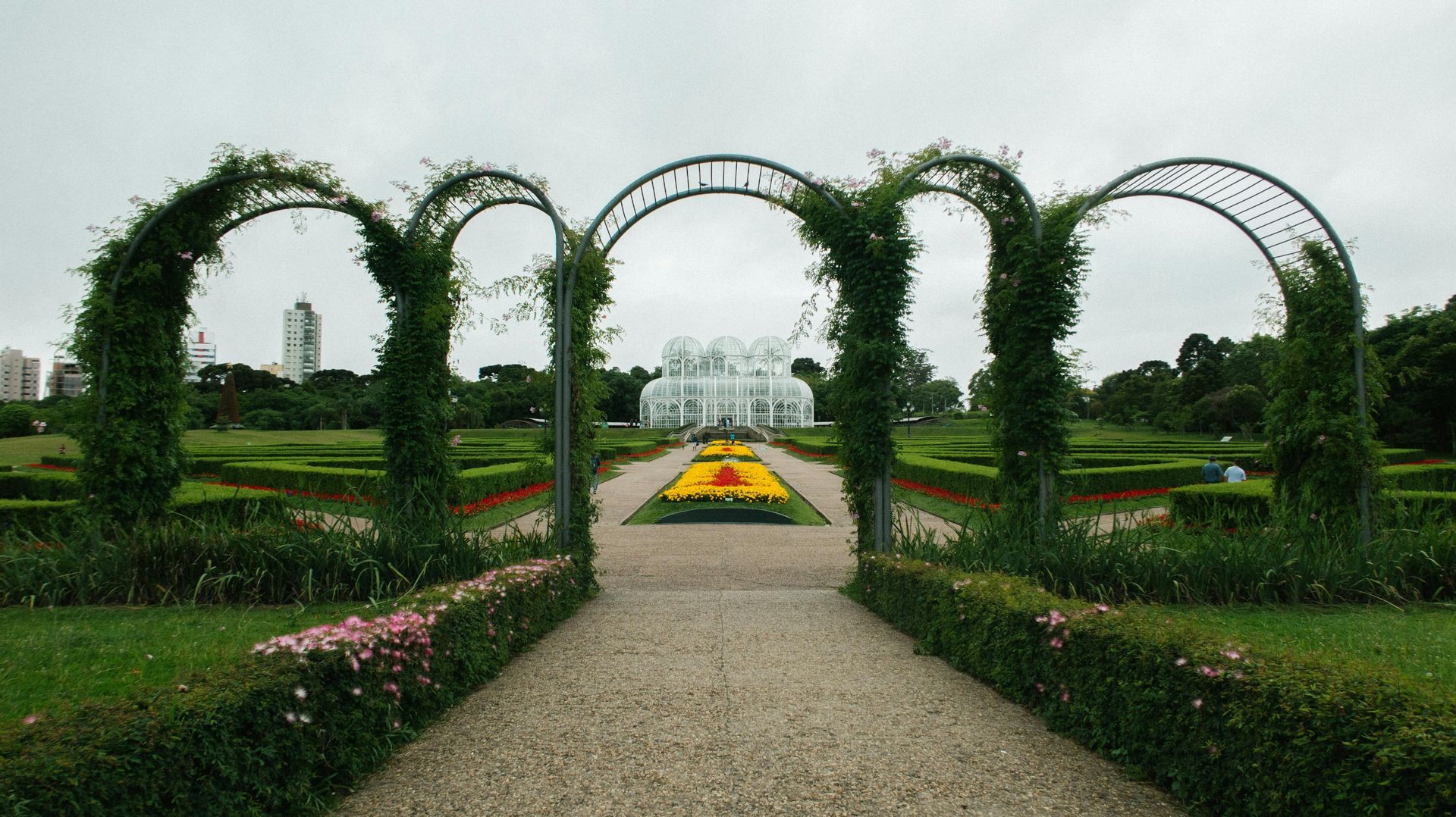 Formal garden with ivy-covered arches framing a gravel path and colorful flower beds under an overcast sky